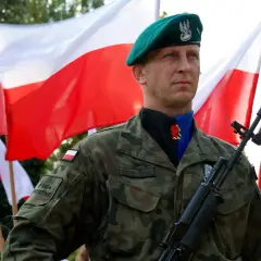Żołnierz w zielonym beretcie, z karabinem i bagnetem, na tle dwóch biało-czerwonych flag.