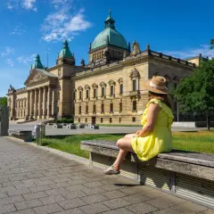 Kobieta w słomkowym kapeluszu i żółtej sukience siedzi na ławce przed monumentalnym budynkiem w Lipsku, mieście w Saksonii znanym z międzynarodowych targów.