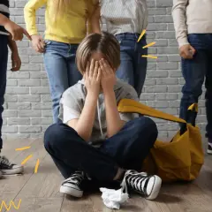 Chłopiec siedzi na podłodze, zakrywając twarz dłońmi, podczas gdy inne dzieci wskazują na niego. To obraz tego, czym jest bullying.