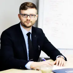 Mężczyzna w garniturze i okularach siedzi przy biurku, pisząc coś na kartce. Obok stoi filiżanka kawy.
