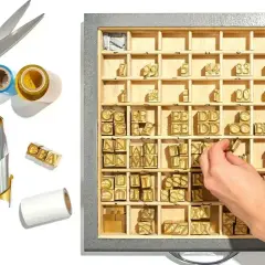 A hand selects brass letter stamps from a tray, ready to create a monogram. Gold scissors and foil rolls are nearby.