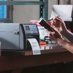 Frau bedient Drucker für Etiketten und Handscanner. Dies ermöglicht effizientes belegloses kommissionieren im Lager.