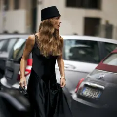 Mujer con sombrero negro y vestido de seda, su pelo en pausa, camina entre coches aparcados.