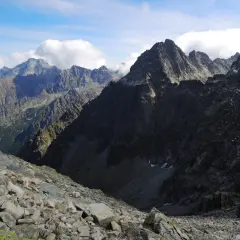 Kamienista ścieżka prowadzi w dół ku skalistym zboczom. W oddali widać ostre szczyty Tatr, w tym Gerlach, a nad nimi kłębiące się chmury.