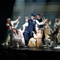 Ensemblen i en svensk revy, med en polisman i centrum, gestikulerar dramatiskt på scenen framför en tågvagnsbakgrund.