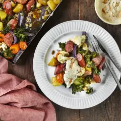 En tallrik med ugnsrostade grönsaker och korv, ett perfekt potatis recept. Bredvid syns en ugnsplåt med samma läckra innehåll.