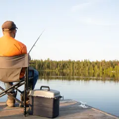Mężczyzna w pomarańczowej koszulce siedzi na krześle wędkarskim na pomoście, łowiąc ryby.