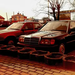 Dwa klasyczne Mercedesy 190e, jeden czerwony, drugi ciemny, stoją obok siebie na brukowanym placu.