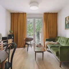 Miodowe zasłony aranżacje tworzą ciepły klimat w salonie. Zielona kanapa i drewniane meble dopełniają przytulną przestrzeń.