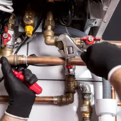 Ręce w rękawiczkach montują instalację gazową w domu. Widoczny schemat połączeń miedzianych rur i zaworów.