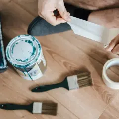 Ręce odrywają taśmę malarską. Obok puszka farby, pędzle i rolka taśmy. Jak usunąć klej po taśmie z drewna?