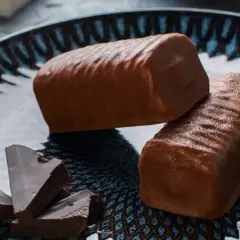 Dwa czekoladowe batony proteinowe na talerzu, obok kawałki gorzkiej czekolady. Idealne do domowego batony proteinowe przepis.