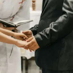 Mani che si stringono durante la dichiarazione matrimonio, con un libro aperto e un sacerdote sullo sfondo.
