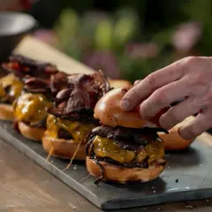 En hand lägger på toppen av en smashed burger med ost och bacon. Flera likadana burgare står uppradade.