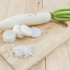 Ein weißer Rettich liegt auf einem Holzbrett, daneben liegen Scheiben und geraspelte Stücke. Perfekt, um Rettich zu fermentieren.
