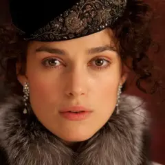 Anna Karenina w eleganckim futrze i ozdobnym kapeluszu, z błyszczącymi kolczykami, patrzy wprost przed siebie.