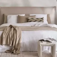Ein gemütliches Schlafzimmer im shabby stil mit einem weißen Bett, beige Decke und Kissen. Ein Beistelltisch mit Lampe und Deko.