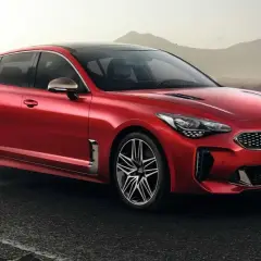 Kia Stinger CV rojo brillante, con llantas deportivas, aparcado en una carretera mojada con montañas al fondo.