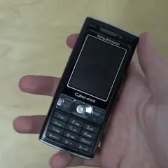 Dłoń trzyma czarny telefon Sony Ericsson k800i z napisem Cyber-shot.