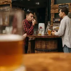 Dwóch mężczyzn rozmawia przy barze, na którym stoi kufel i szklanka piwa. Dyskutują, ile schodzi jedno piwo, podczas gdy w tle widać drewniane dekoracje i plakaty.