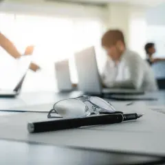 Okulary i długopis na dokumentach. W tle ludzie pracujący przy laptopach, być może praca na czarno.