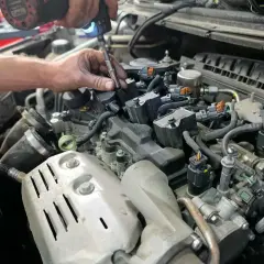 Mécanicien utilisant une visseuse pour changer les bougies d'allumage essence. Un moteur de voiture est visible.