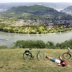 Pareja descansa junto a sus bicicletas en la ruta EuroVelo 15, con un río y un pueblo en el valle.