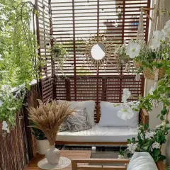 Przytulny balkon w bloku z drewnianymi zasłonami, sofą, białymi kwiatami i makramami.