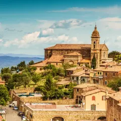 Panoramic view of Montalcino, Italy, with terracotta roofs, a church, and rolling hills under a blue sky.