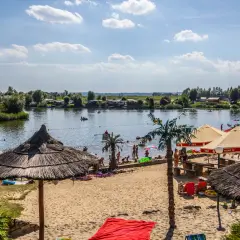 Słoneczny dzień na plaży w Kuźni Raciborskiej. Ludzie odpoczywają pod parasolami, kąpią się w jeziorze i pływają łódkami.