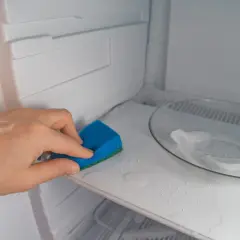 Mano limpiando el interior de una nevera con una esponja azul. El estante está mojado y hay un plato con hielo.