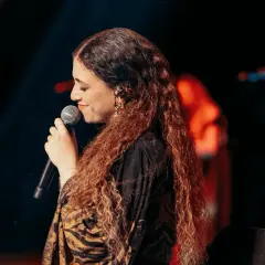 Cantante con lunghi capelli ricci, microfono in mano, sul palco. Un'espressione che evoca la bellezza della Sicilia, un testo che risuona.
