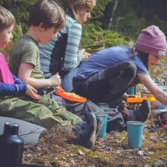 Familj njuter av en picknick i skogen, kanske vid restaurang Ånnaboda. Barn äter pasta och vuxna förbereder mat.