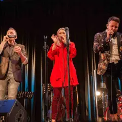Trzech wokalistów na scenie podczas warsztatów wokalnych. Kobieta w czerwieni, mężczyzna w marynarce z wzorami i mężczyzna grający na harmonijce.