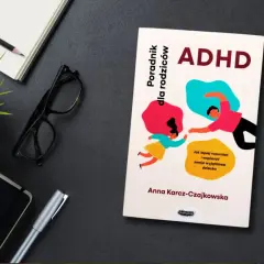 Książka "ADHD. Poradnik dla rodziców" Anny Karcz-Czajkowskiej, z ilustracją rodzica i dziecka. Obok notatnik, okulary i długopis.