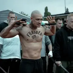 Wytatuowany mężczyzna z napisem "Furioza" na klatce piersiowej, otoczony przez grupę mężczyzn, przygotowuje się do konfrontacji.