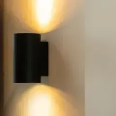 Lámpara de pared original negra, proyectando luz cálida hacia arriba y abajo. Copas de cristal en un armario a la izquierda.