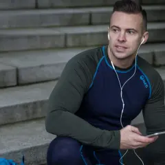 Mężczyzna w zielonej koszulce termoaktywnej z granatowymi rękawami i niebieskimi szwami, słucha muzyki na telefonie, siedząc na schodach.
