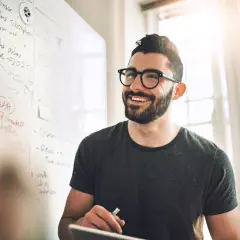 Ein Mann mit Brille lächelt vor einer Tafel mit Notizen zur Fernuni Wirtschaftsinformatik.