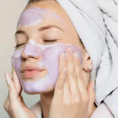 En kvinna med handduk i håret applicerar en lila ansiktsmask. En avkopplande ansiktsbehandling hemma för att skämma bort sig själv.