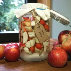 Ein Glas mit Apfelstücken und der Aufschrift "Apfel-essig" steht neben frischen Äpfeln. Manche glauben, Apfelessig hilft gegen Cholesterin.