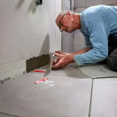 Man limma kakel på golvet, noggrant placerar han en ny platta. Kakelkryss och kilar syns på golvet.