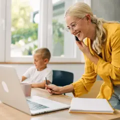 Uśmiechnięta kobieta rozmawia przez telefon, pracując przy laptopie, podczas gdy jej syn rysuje obok. Odpowiedź na pytanie: czy na urlopie wychowawczym można pracować.