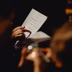 Mani che seguono le note di una canzone funerale su uno spartito, in un'atmosfera raccolta e commossa.