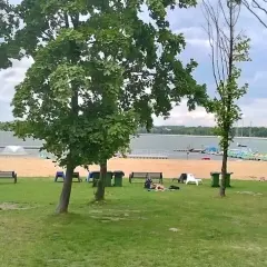 Zielone **łąka** nad **jeziorem**, piaszczysta plaża z ławkami i odpoczywającymi ludźmi, w tle łodzie i drzewa.