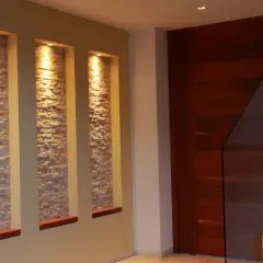 Tre nicchie in cartongesso illuminate da faretti, con rivestimento in pietra, creano un punto focale moderno accanto a una porta in legno e una scala con parapetto in vetro.