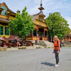 Kolorowe, drewniane budynki w Czechach, pełne ciekawostek dla dzieci. Kobieta w pomarańczowej bluzce pozuje na tle malowniczej architektury.