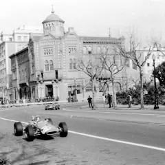 Coches de carreras compiten en el circuito de Montjuïc, con espectadores en las gradas y edificios históricos al fondo.
