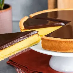 Una fetta di torta Susanna, con crema gialla e copertura al cioccolato fondente, viene sollevata da una tortiera su un'alzata bianca.