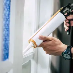 Ręce trzymają pistolet do silikonu, gotowe do uszczelnienia okna. Dowiedz się, jak otworzyć silikon i wykonać pracę.